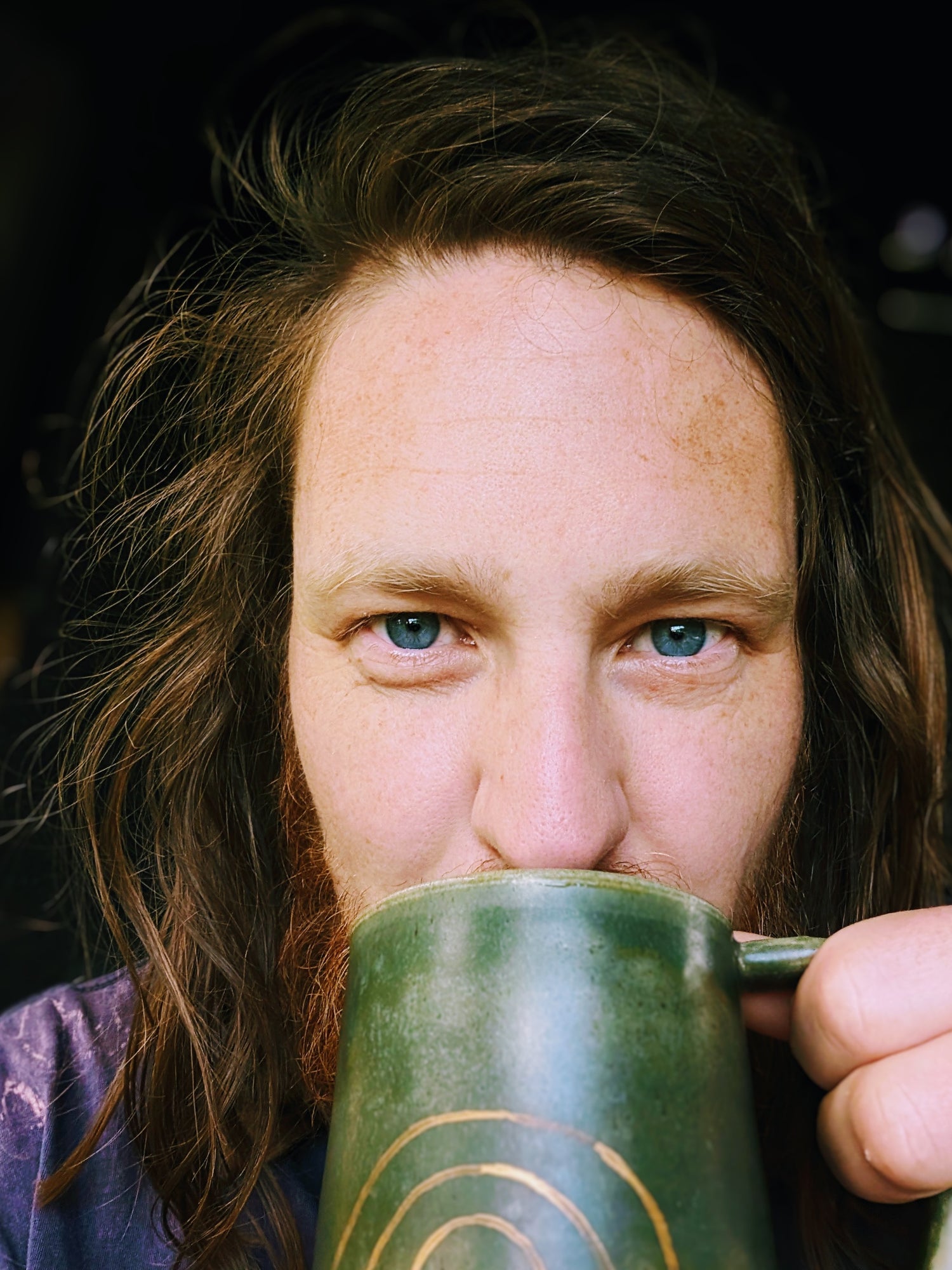 Person with long hair holding a green mug close to their face against a dark background